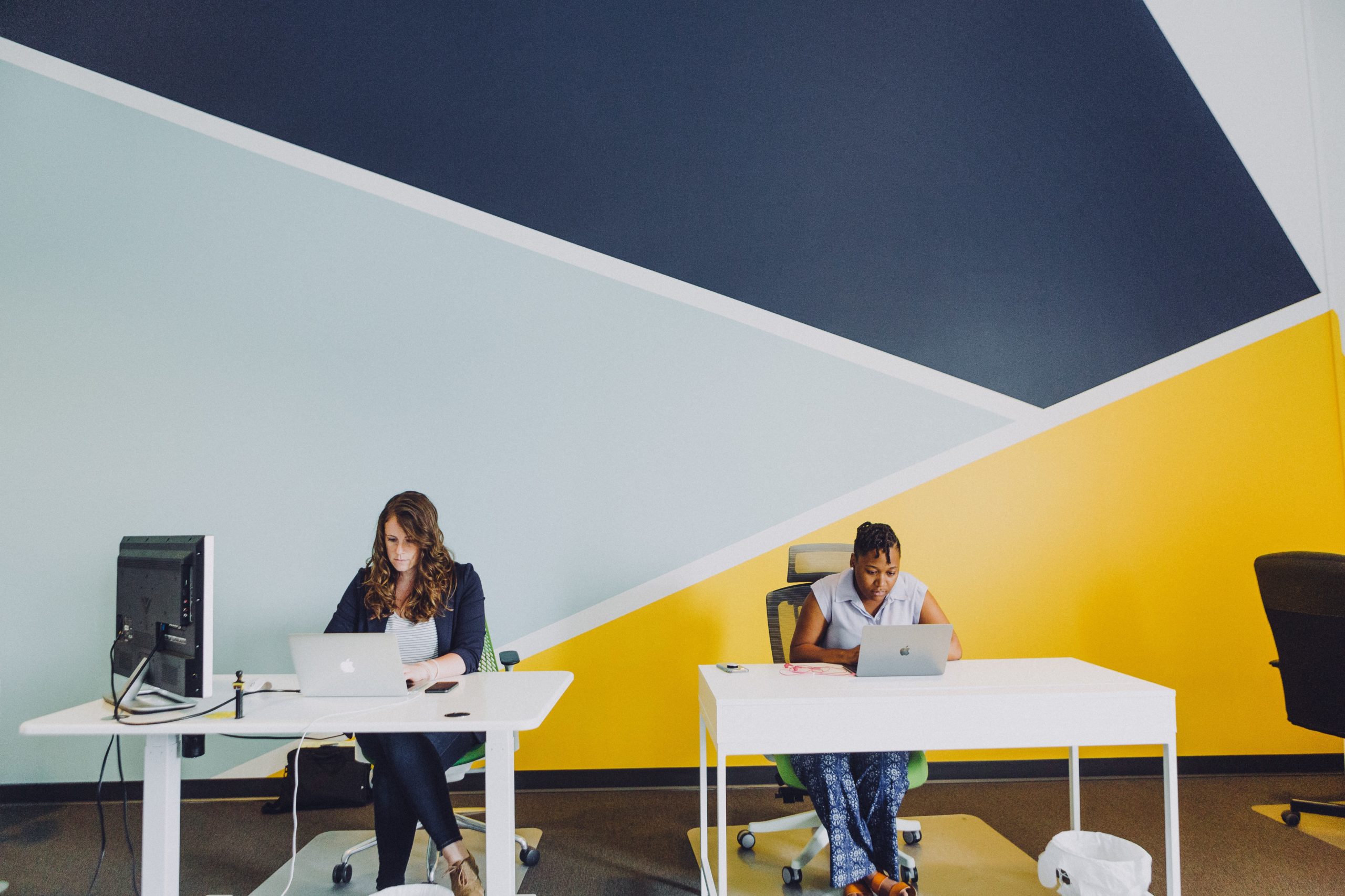 White woman and black woman sitting at desk working at a laptop with a blue and yellow geometric pattern on the wall behind them.